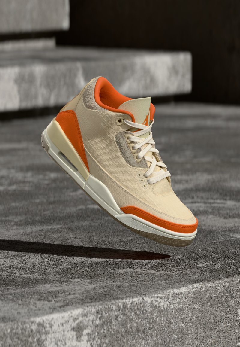 Beige og lys orange sneaker med hvide snørebånd, mellemsål med luftpolstring og struktureret overdel, hængende over grå betonstrin.