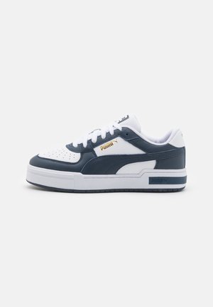 Baskets blanches et navy en cuir lisse avec un bout perforé, arborant un logo doré sur le côté et une semelle blanche épaisse.