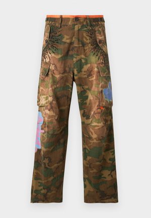 Homme portant une casquette rouge, une veste en jean bleu délavé sur une chemise blanche, un pantalon cargo camouflage avec des accents roses, et des chaussures plateformes noires, debout droit.