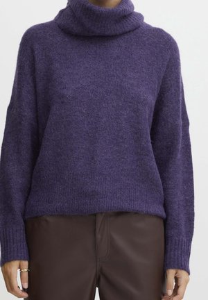 Paarse, zachte coltrui gedragen met donkerbruine broek, toont bovenlichaam en armen tegen een effen achtergrond.