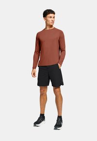 T-shirt de sport à manches longues couleur rouille avec un tissu évacuant l'humidité, associé à un short noir. Chaussures de course noires avec des semelles blanches.