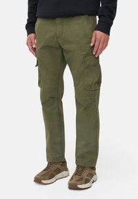 Olivgrüne Cargo-Hosen mit Seitentaschen, aus einem robusten Stoff gefertigt. Sie haben eine gerade Passform und verstärkte Nähte. Kombiniert mit braunen Sneakers.