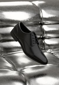 Zapato de vestir de cuero negro con acabado pulido, que presenta una puntera redondeada, diseño con cordones y sutiles acentos de costura, flotando sobre una superficie brillante.