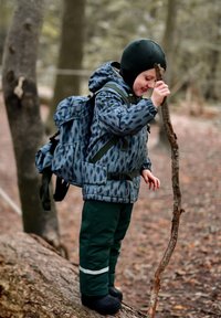 Barn iført en blå mønstret jakke og grønne bukser, stående på en træstamme og holder en pind i et skovområde. Sorte støvler og rygsæk er synlige.