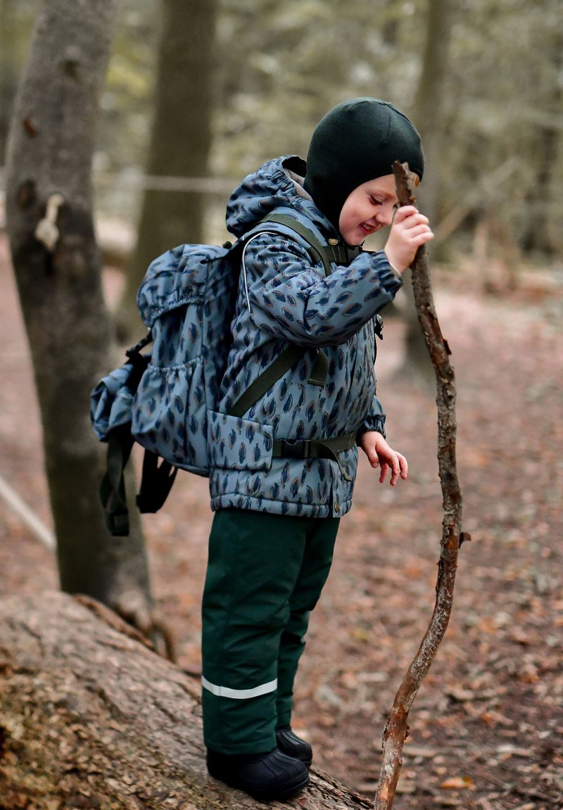 Barn iført en blå mønstret jakke og grønne bukser, stående på en træstamme og holder en pind i et skovområde. Sorte støvler og rygsæk er synlige.