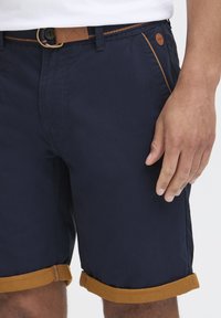 Marineblå shorts med rullet sennepsfarget kant, sidelommer og et brunt belte med metallspenne. Minimalistisk design med subtile sømdetaljer.