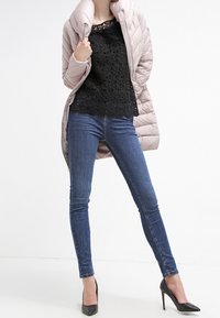 Cappotto imbottito ripiegabile di colore rosa chiaro, top in pizzo nero, jeans skinny blu e tacchi a punta neri, caratterizzato da un design trapuntato e collo arricciato.