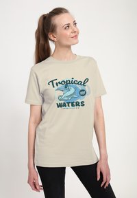 Béžové bavlněné tričko s grafickým potiskem žraloka kladivoun a textem "Tropické vody" v tmavě zelené a modré.