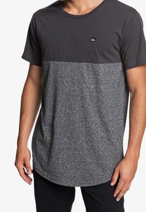 Homme portant un T-shirt bicolore gris avec une partie supérieure noire et une partie inférieure gris chiné, debout les bras détendus le long du corps.