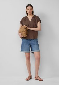 Blouse marron à manches courtes avec un col en V, shorts en denim avec des poignets retroussés et sandales marron ; tenant un sac en osier beige.