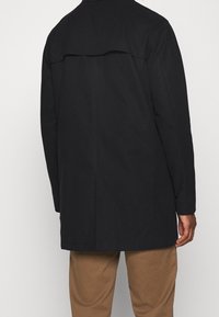 Svart, medellång kappa med en knappförsedd bakflik. Har en slät textur och skräddarsydd passform, i kombination med khakifärgade byxor.