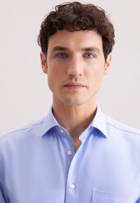 Lichtblauw, overhemd met lange mouwen en een button-down kraag, voorzak en een gladde stoftextuur. Hoofd en schouders van het model zichtbaar.