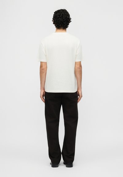 Άνδρας με σγουρά μαλλιά στέκεται γυρισμένος με την πλάτη, φοράει απλό λευκό μπλουζάκι, μαύρο παντελόνι και μαύρα παπούτσια μπροστά σε απλό φόντο.