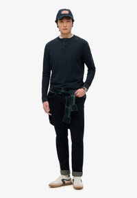 Maglietta henley a maniche lunghe blu navy, jeans scuri, camicia a scacchi verde legata in vita, scarpe bianche con dettagli neri e un cappellino blu navy con una toppa a forma di bandiera.