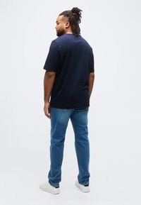 Άνδρας με dreadlocks στέκεται με την πλάτη, φοράει μπλε σκούρο μπλουζάκι, μπλε τζιν και λευκά αθλητικά παπούτσια μπροστά σε ένα απλό φόντο.