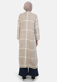 Long cardigan beige avec motif à carreaux blancs, deux poches latérales et manches longues ; fabriqué en tissu texturé ; porté sur un pantalon noir à jambes larges.