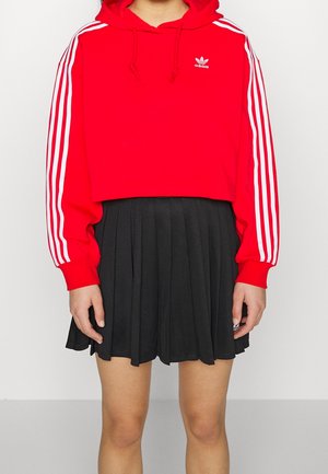 Osoba nosząca czerwoną krótką bluzę Adidas z białymi paskami na rękawach i czarną plisowaną spódnicę, stojąca na białym tle.