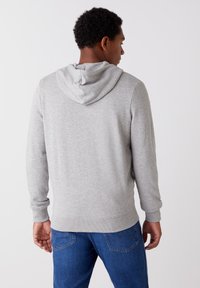 Szara bluza z kapturem z miękkiego materiału, z troczkiem, ściągaczami przy rękawach i dole, noszona z niebieskimi dżinsami, ukazująca luźny krój z tyłu.