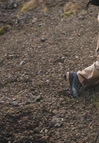 Brown hiking pants, gray and blue trail shoes with textured soles, walking on rocky terrain. Ground features small stones and gravel.