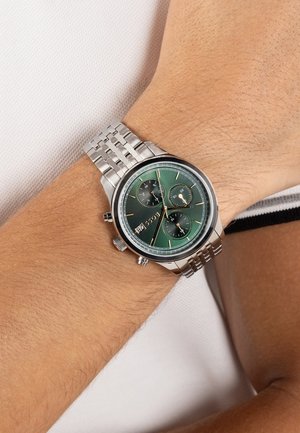 Silver metal wristwatch with green dial, three subdials, date window, and chronograph buttons worn on left wrist of person in white garment.