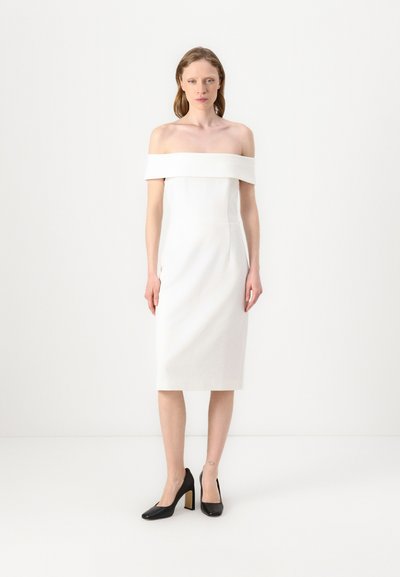 Hvid off-shoulder kjole med en tætsiddende silhuet, knælængde og glat stof. Båret med sorte højhælede sko.