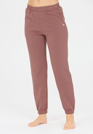 Femme portant un pantalon de jogging mauve à taille élastique avec des poignets élastiques, debout pieds nus sur un fond uni.
