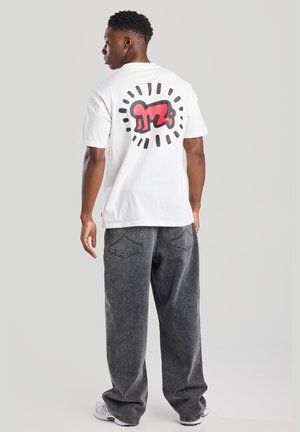 White short-sleeve t-shirt featuring a large red and black graphic on the back, paired with gray baggy jeans and white sneakers.