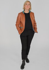 Brun läderjacka med turkosa detaljer, svart figurnära topp och svarta byxor. Modellen bär svarta ankelstövlar och står självsäkert.