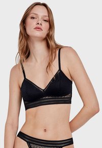 Bralette en dentelle noire avec des fines bretelles réglables, un décolleté en V et un motif texturé. Présente une bande en dentelle transparente en bas.