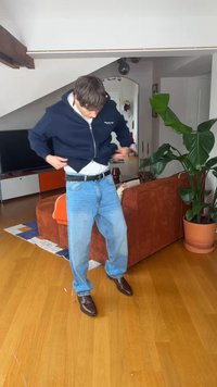 Moški v zaprtih prostorih, oblečen v modro kapucarico, svetlo modre kavbojke in rjave čevlje, prilagaja svojo obleko blizu rjavega kavča in velike lončnice.