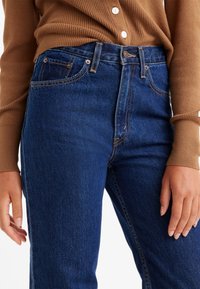 Jeans de mezclilla azul de cintura alta, con bolsillos frontales, costuras en contraste y una textura suave. Combinados con un suéter marrón de canalé.