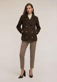 Ruskea kaksirivinen villatakki, jossa suuret napit ja kaksi etutaskua, puettu kermaan vivahtavan pooloneuleen päälle, yhdistetty kuvioituihin housuihin ja korkokenkiin.