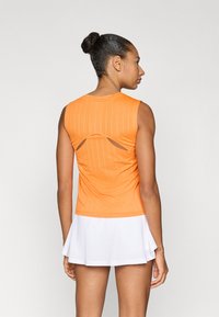 Πορτοκαλί αθλητικό τοπ χωρίς μανίκια με κάθετες ρίγες, που διαθέτει άνοιγμα στην πλάτη. Συνδυάζεται με λευκή φούστα-σορτς με καμπάνα.