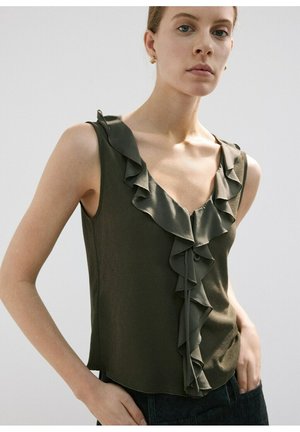 Femme portant un haut sans manches vert foncé avec un col en V à volants et un détail noué à l'avant, posant avec la main dans la poche devant un fond uni.