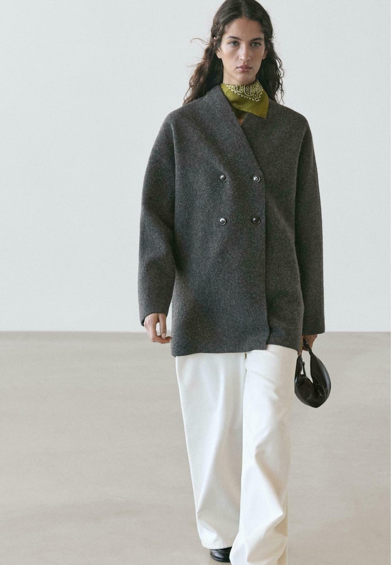 Cappotto grigio doppiopetto in lana con grandi bottoni, indossato sopra una sciarpa verde, abbinato a pantaloni bianchi a gamba larga, con una borsa nera in mano.