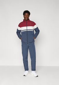 Vindjakke og joggersæt i marineblå, burgundy og hvid. Jakken har lynlåslukning, høj krave og logo. Glat, letvægtsstof.