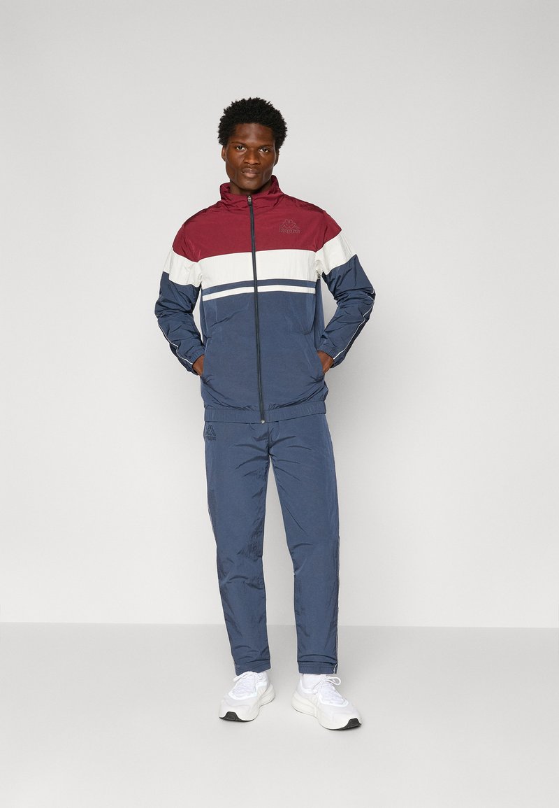 Windbreaker- und Jogginganzugset in Marineblau, Burgunderrot und Weiß. Die Jacke hat einen Reißverschluss, einen hohen Kragen und ein Logo. Glattes, leichtes Material.