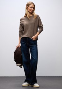 Brązowy sweter dzianinowy, luźny krój, dekolt w kształcie litery V; ciemnoniebieskie spodnie typu wide-leg; trzyma ciemnobrązową kurtkę bomber; białe sneakersy; gładkie faktury.