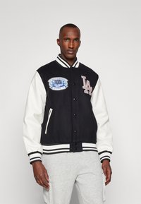 Zwart-witte varsityjack met een wollen lichaam, leren mouwen, geribde manchetten en een geborduurd logopatch op de borst.