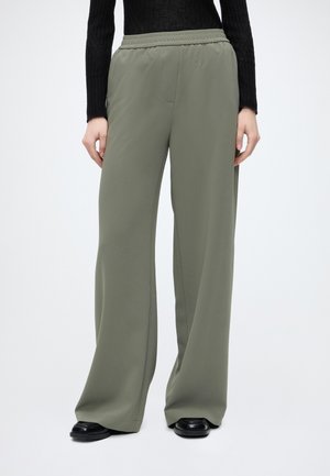 Personne portant un pantalon large ample vert olive avec une taille élastique et des chaussures noires, debout devant un fond clair uni.