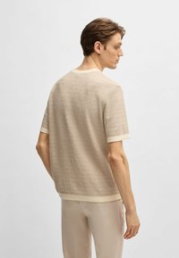 Maglione corto a maniche corte beige con una scollatura e orlo a coste color crema, indossato con pantaloni chiari, visto da dietro.