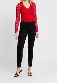 Röd spets långärmad bodysuit med djup V-ringning, knapprad, i kombination med höga svarta skinny jeans och svarta remsandaler.