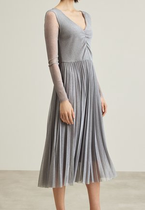 Femme portant une robe grise à manches longues avec un haut froncé à col en V et une jupe plissée, debout devant un fond clair uni.