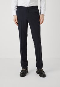 Pantaloni da dress blu navy con una vestibilità sartoriale, caratterizzati da una texture liscia, tasche laterali e doppi bottoni in vita. Abbinati a mocassini neri.