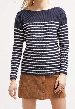 Personne portant un pull à manches longues rayé bleu marine et blanc avec une jupe en suède marron à boutons.