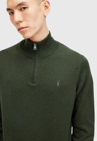 Žalias zip-up megztinis pagamintas iš mezginio medžiagos, su aukštu apykakle, iškilusiu logotipo detalė ir lygiu paviršiumi.