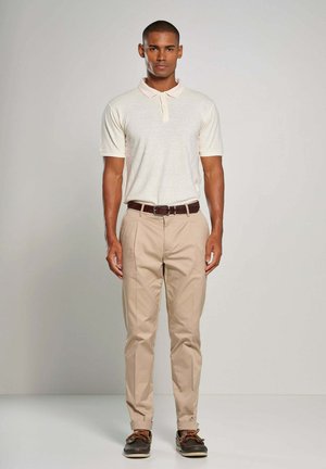 Mężczyzna stoi, ubrany w kremowe polo z krótkim rękawem, beżowe spodnie z brązowym paskiem i ciemne buty typu boat na jednolitym jasnym tle.