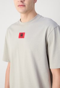 T-shirt en coton gris clair à manches courtes et col rond. Présente un carré rouge avec un texte noir et un logo sur la poitrine.