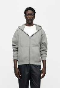 Γκρι φούτερ με φερμουάρ και κουκούλα με κορδόνι, με ριμπ μανσέτες και τελείωμα, φορεμένο πάνω από λευκό πουκάμισο, σε συνδυασμό με σκούρο παντελόνι. Λείο ύφασμα.