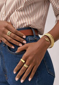 Des bagues et des bracelets en ton or ornent des mains reposant sur une taille en denim. Les bagues présentent des finitions texturées et lisses, avec des largeurs variées.